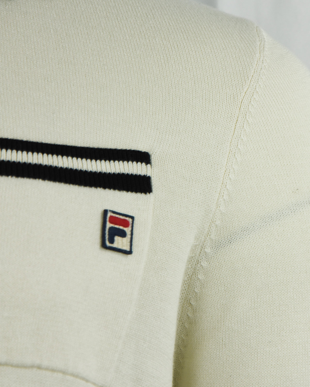Close-up of cream polo pocket with black stripe trim and Fila logo patch.