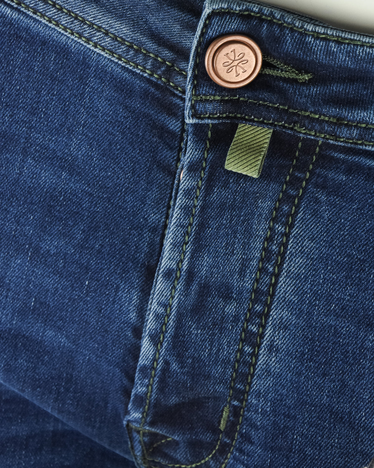 Denim fly area with copper button, green inner label, and contrast seam stitching.