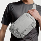 Ash gray sling bag worn crossbody, with front flap and buckled straps