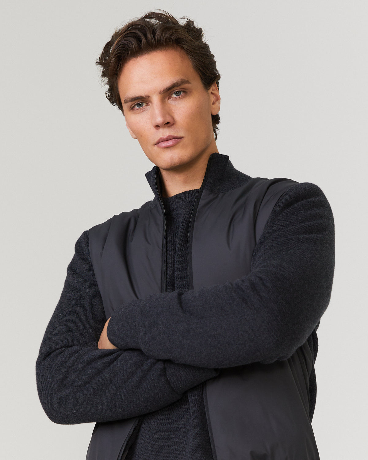 Men's hybrid jacket worn with arms crossed; navy knit sleeves, stand collar, zip front.